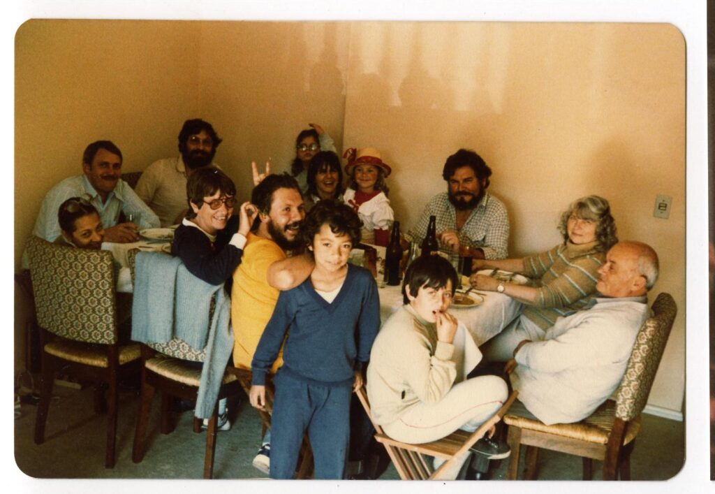 An old family photo of a big family gathered around a lunch table, smiling at the camera.