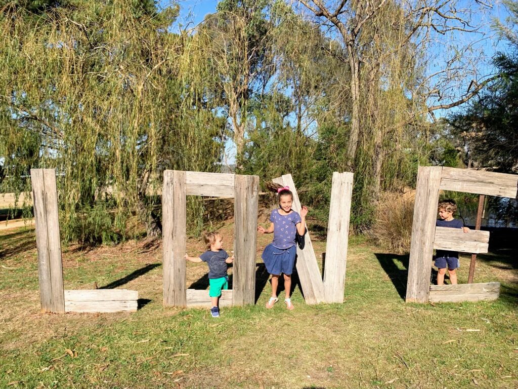 Three children playing among giant wooden letters spelling LOVE in a sunny park.