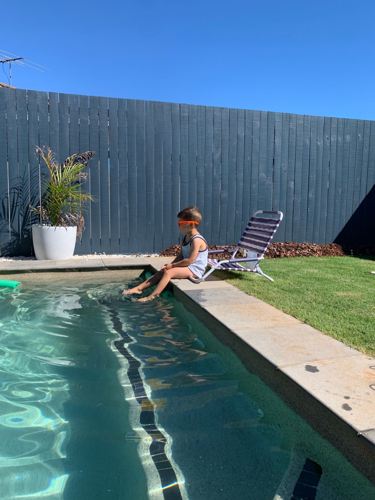 A young boy in orange goggles sits on the edge of a backyard pool, dipping his feet in the water.